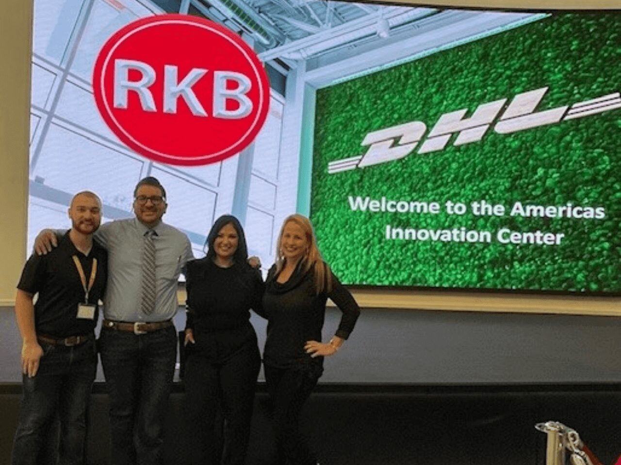 Four people posing in front of RKB and DHL logos at an innovation center.
