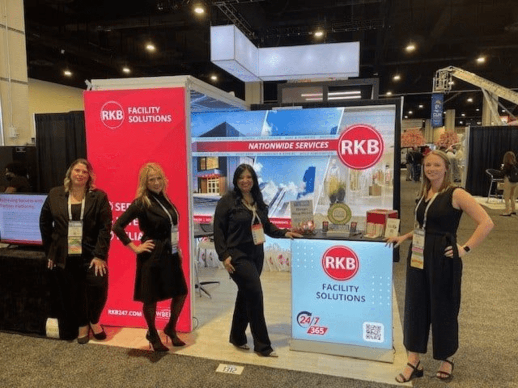 Three women standing in front of an RKB trade show booth with promotional displays.